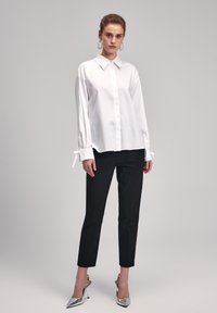 Camicia bianca con bottoni e maniche lunghe, con nastri nei polsini; abbinata a pantaloni neri slim-fit e décolleté argentati a punta.