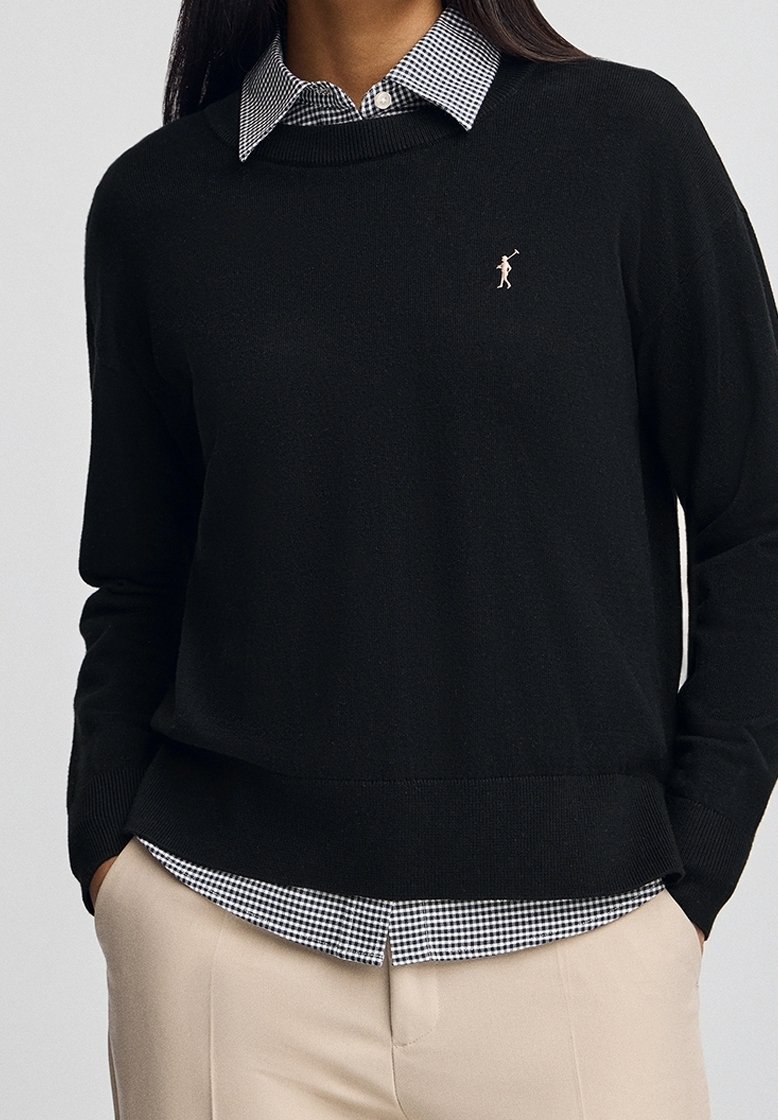 Femme portant un pull noir avec un petit logo de joueur de polo sur une chemise à carreaux noir et blanc, pantalon beige, mains dans les poches.