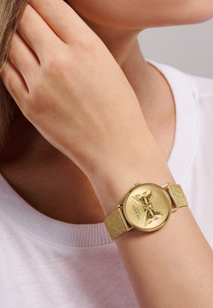 Ceas auriu cu o curea texturată, cadran rotund cu un design în formă de fundă și un cadran minimalistic, purtat la încheietura mâinii.