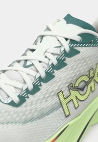 Ukrupnjen prikaz bijele i zelene sportske tenisice s bijelim vezicama i teksturiranim potplatom, s logotipom "HOKA" sa strane.