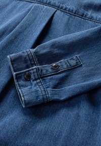 Jeansskjortärmh i mörkblått. Har en knappmanchett med två svarta knappar, strukturerad tyg med en slät yta och subtil lyster.