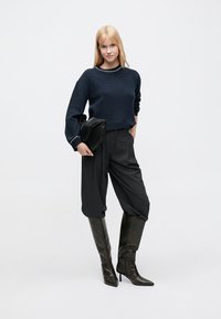 Pull bleu marine en tissu texturé avec un col orné de perles, associé à un pantalon noir taille haute et des bottes en cuir noires montantes jusqu'aux genoux.