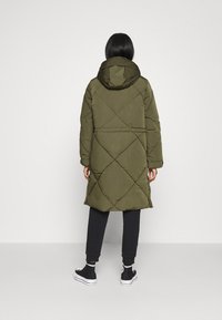 Abrigo largo acolchado color verde oliva con capucha, que presenta un patrón acolchado en forma de diamante y puños elásticos. Lleva puesto sobre pantalones oscuros y zapatillas.