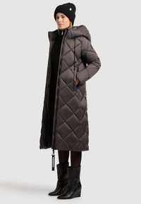 Lange, donkergrijze gewatteerde pufferjas met een capuchon, voorzien van een ritssluiting, koordjes en zijzakken. Draagt over een zwarte outfit en laarzen.