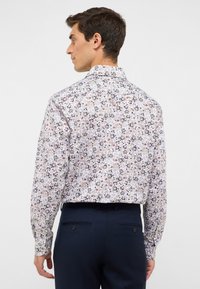 Uomo di spalle con una camicia bianca a maniche lunghe con un motivo floreale marrone e blu, e pantaloni eleganti blu.