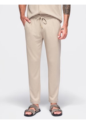 Pantalon beige décontracté avec cordon de serrage, porté avec des sandales taupe à double bride, montrant des avant-bras et des mains tatoués en pose détendue.