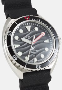 Montre Fossil avec un bracelet en caoutchouc noir, boîtier en acier inoxydable argenté, cadran gris texturé, marqueurs lumineux, accent rouge sur la seconde.