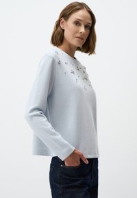 Hellblauer Pullover mit langen Ärmeln, der auf der Vorderseite mit facettierten Edelsteinakzenten versehen ist, aus weichem Stoff mit lockerer Passform.