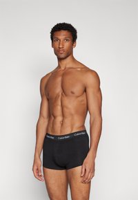 Schwarze Baumwoll-Boxershorts mit einem elastischen Bund mit Markenlogo. Das Modell hat einen muskulösen Körperbau und steht vor einem schlichten grauen Hintergrund.