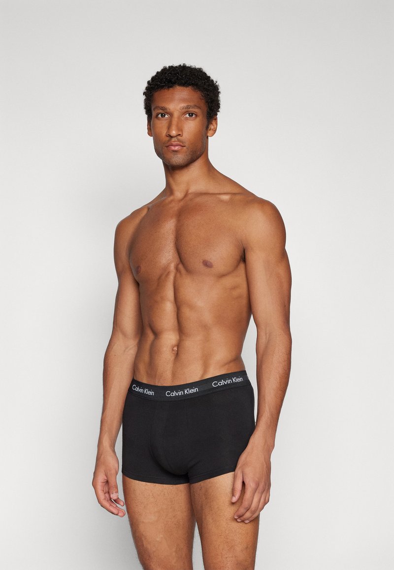 Schwarze Baumwoll-Boxershorts mit einem elastischen Bund mit Markenlogo. Das Modell hat einen muskulösen Körperbau und steht vor einem schlichten grauen Hintergrund.