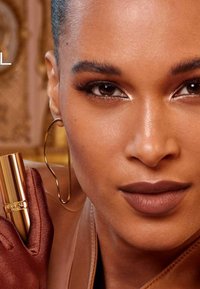 Rossetto marrone in un tubetto dorato, tenuto da una mano guantata. La modella ha un finish lucido sulla pelle e grandi orecchini a cerchio dorati.