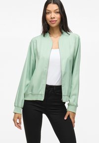 Giacca bomber verde chiaro realizzata in morbido tessuto satinato, con chiusura a zip e polsini a coste, abbinata a una maglietta bianca e jeans neri.