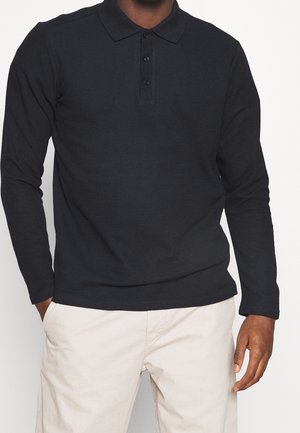 Man in een zwart poloshirt met lange mouwen en lichtbeige broek, staande met één hand in zijn zak tegen een effen achtergrond.