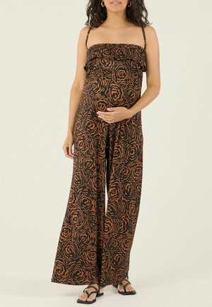 Schwangere Frau in einem braun-schwarzen, floralen trägerlosen Jumpsuit und schwarzen Sandalen, die eine Hand auf ihrem Babybauch ruhen lässt.
