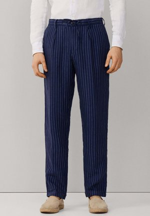 Homme portant un pantalon sur mesure à rayures bleu marine, une chemise blanche et des espadrilles beiges, debout devant un fond uni.