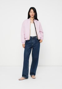 Femme portant une veste bomber violette claire, un haut blanc, un pantalon bleu à large jambe et des ballerines roses claires, posant sur un fond blanc uni.