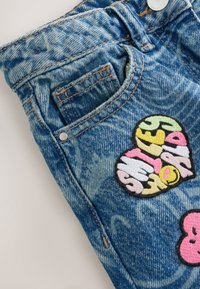 Poche de jean en denim bleu avec des patchs colorés en forme de cœur, un patch a un visage souriant jaune et un design de texte multicolore.