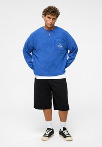 Blauwe, oversized sweatshirt met een voorzak en witte geborduurde tekst, gecombineerd met zwarte shorts en sneakers, met zichtbare witte sokken.