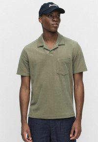 Oliivivihreä lyhythihainen polo-paita, jossa on teksturoitu pinta, litteä kaulus ja rintatasku; yhdistettynä tummansiniseen lippikseen, jossa on valkoiset kirjaimet.