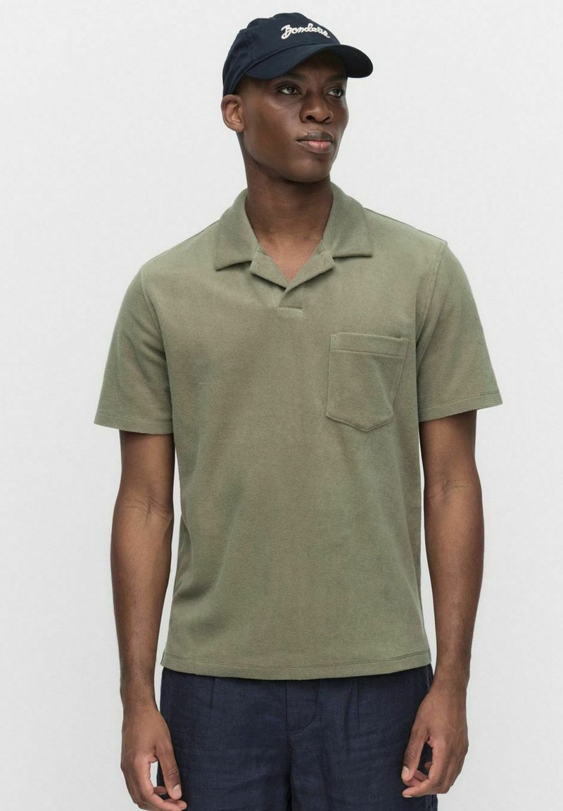 Oliivivihreä lyhythihainen polo-paita, jossa on teksturoitu pinta, litteä kaulus ja rintatasku; yhdistettynä tummansiniseen lippikseen, jossa on valkoiset kirjaimet.