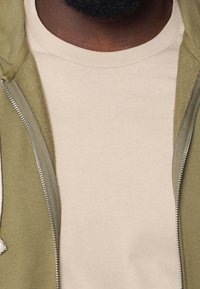 Primer plano de una persona que lleva una camiseta de cuello redondo beige debajo de una chaqueta de cremallera verde oliva abierta.