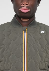 Többrétegű olajzöld kabát magas gallérral, elől cipzárral, sárga és fehér csíkokkal, valamint logóval a mellkason.