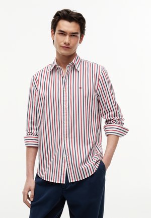 Jeune homme portant une chemise blanche à rayures verticales rouges et bleues et un pantalon bleu, debout avec une main dans la poche.