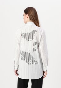 White button-up shirt with vertical black stripes, featuring a contrasting black graphic design on the back. Long sleeves and tailored fit.