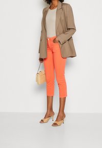 Kobieta w dopasowanej beżowej marynarce nad jasnoszarym topem, w zestawieniu z dopasowanymi koralowymi spodniami 7/8 i beżowymi sandałami na obcasie, trzymająca plecioną torbę.