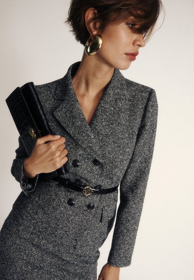 Blazer gris texturé avec des boutons double poitrine, taille ceinturée noire et une pochette noire lisse. Des boucles d'oreilles circulaires dorées complètent le look.