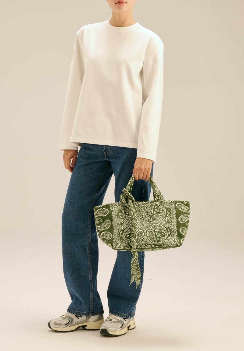 Zielona torba w wzór paisley z materiałowymi wiązaniami, trzymana przez osobę w białej bluzce z długim rękawem i niebieskich dżinsach, noszącą szare sneakersy.