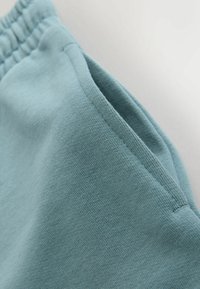 Svetlomodrá bavlnená tkanina s viditeľným prešívaním a časťou elastického pásu, zobrazujúca otvor bočného vrecka.