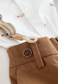 White button-up shirt with small plane prints, paired with brown fabric pants featuring a stitched waistband and a round brown button.
