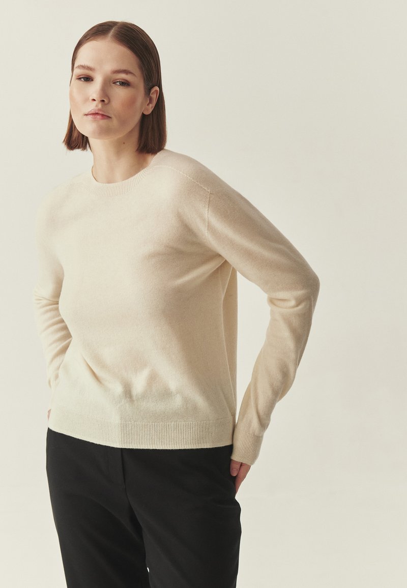 Pull beige en maille douce avec un col rond et des manches longues. Le tissu présente une texture lisse, associé à un pantalon noir.
