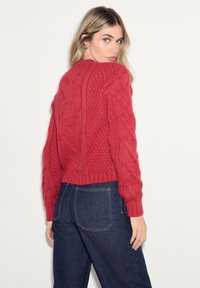 Red knitted sweater with a cable knit pattern and ribbed cuffs, paired with dark blue denim jeans. Styling is relaxed and casual.