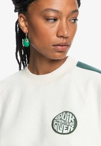 Suéter crema con acentos verdes, con un logo bordado redondo. La tela parece texturizada con un diseño de cuello redondo acanalado.