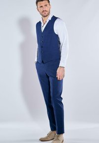 Marineblauer dreiteiliger Anzug mit einer taillierten Weste und maßgeschneiderten Hosen, kombiniert mit einem weißen Hemd und beigen Schuhen. Glatte Stoffstruktur.