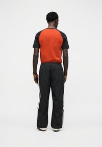 Άνδρας που φοράει κόκκινο και μαύρο κοντομάνικο πουκάμισο και μαύρο παντελόνι φόρμας με λευκές ρίγες στα πλάγια, στέκεται με την πλάτη γυρισμένη σε απλό λευκό φόντο.