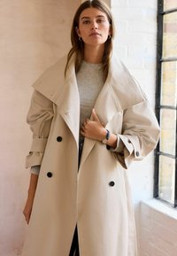 Beige trenchcoat med en stor krave, dobbeltknapning, sorte knapper og opkrøllede ærmer; båret over en grå top.