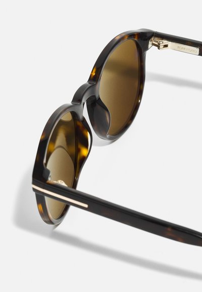 Ochelari de soare cu model țestoasă, cu lentile maro, formă rotundă și accente metalice. Prezentând o decupare unică în jurul lentilelor pentru un detaliu de design.