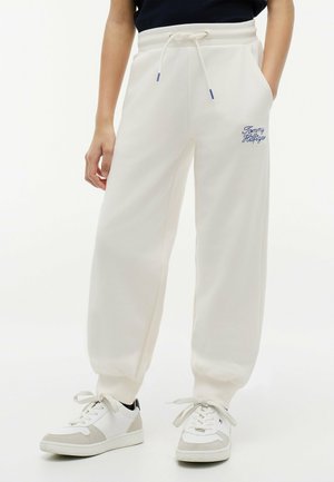 Persona che indossa pantaloni della tuta bianchi Tommy Hilfiger con coulisse in vita e sneakers bianche, in piedi davanti a uno sfondo semplice.