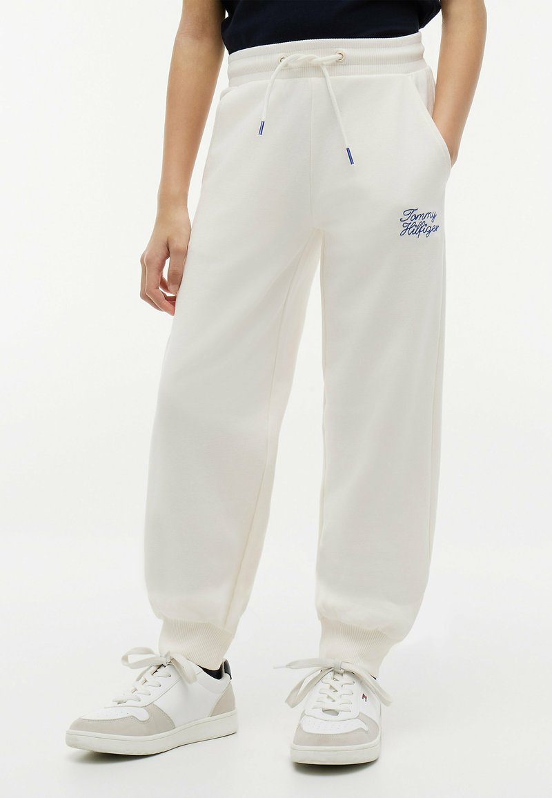 Personne portant un pantalon de jogging blanc Tommy Hilfiger avec taille à cordon et des baskets blanches, debout devant un fond uni.