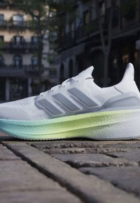 Baskets blanches de sport avec semelle dégradée bleu clair et verte, placées sur une rue pavée avec des bâtiments flous en arrière-plan.