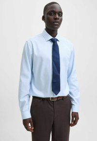 Lichtblauw overhemd met lange mouwen en een subtiel patroon, gecombineerd met een donkerblauwe stropdas. Bruine riem complementeert donkerbruine broek.