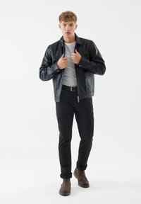 Giacca di pelle nera con design con colletto, chiusura con zip e due tasche frontali. Indossata sopra una camicia bianca e abbinata a pantaloni neri e stivali marroni.