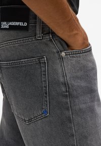 Sivé džíny z denimu s jemnou textúrou, s čiernou nášivkou na páse, modrým kontrastným prešívaním a štandardnými zadnými vreckami.