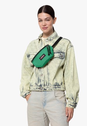 Joven mujer vistiendo una chaqueta de mezclilla deslavada y jeans, llevando un bolso cruzado verde con una correa negra y un logo en el pecho.