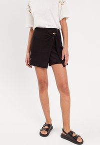 Shorts noirs en tissu texturé, dotés d'un détail de nœud sur le côté et d'un ourlet légèrement arrondi, associés à des sandales noires.