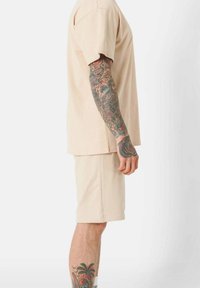 Beige cotton t-shirt and shorts set, short sleeves, relaxed fit, smooth texture, with intricate tattoos visible on forearm.