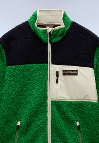 Fleece dzseki magas gallérral, zöld, sötétkék és krém színű panelekből, texturált felülettel és logóval ellátott cipzáras mellzsebbel.
